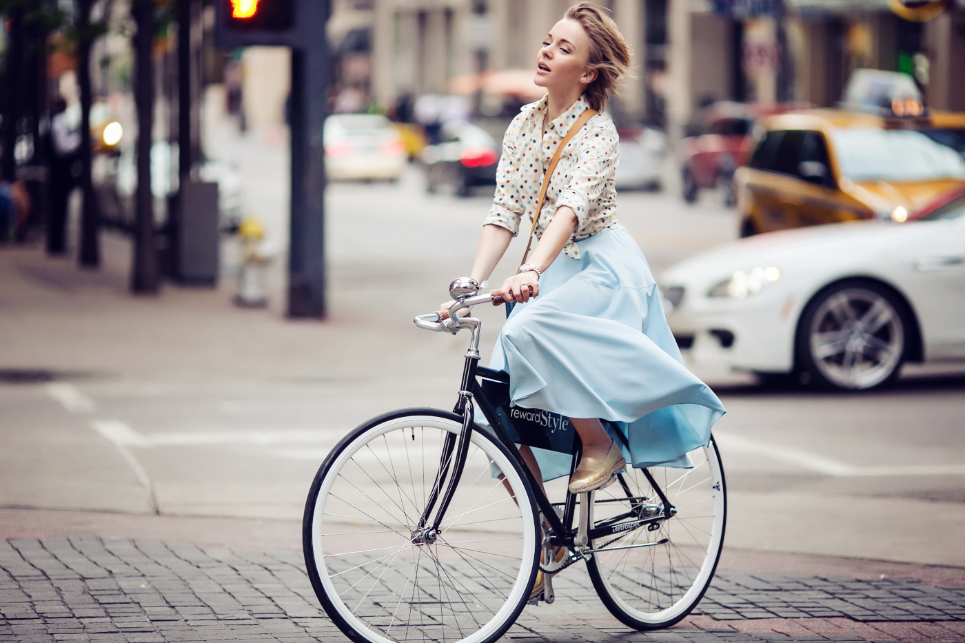 darya kamalova a fashion blogger from thecablook in dallas texas on the bycicle in polka dot shirt and blue skirt with gold espadrilles and other storie bag for rewardstyle rsthecon